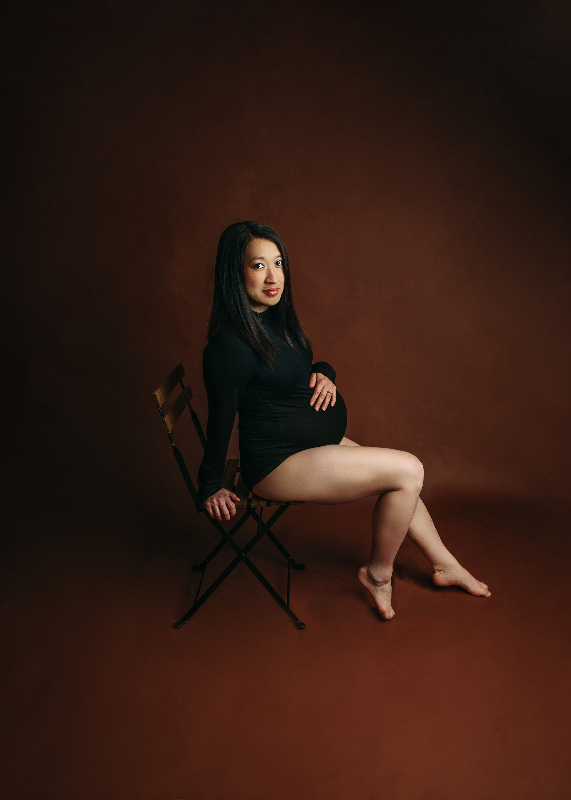 maternity in black bodysuit on chair and chestnut backdrop minneapolis maternity photography
